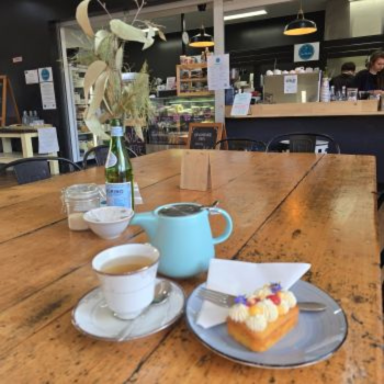 Wooden café table set with a teapot, cup of tea and a decorated pastry, with the café counter visible in the background.
